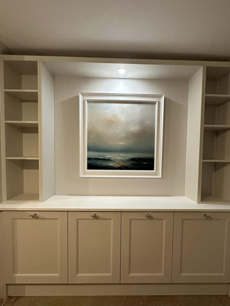 Head on image of a furniture wall with white units and an LED light in the top lighting a painting situated in the middle of the wall. The painting is of a cloudy sky over the sea.